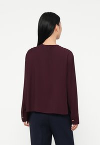Woman with long black hair wearing a long-sleeve dark purple blouse and dark pants, shown from the back against a white background.