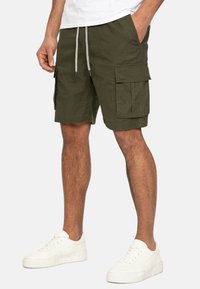 Shorts cargo vert olive avec deux poches latérales et deux poches avant à rabat. Fabriqués en tissu texturé, avec une taille réglable par cordon et des baskets blanches.