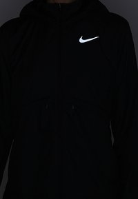 Veste coupe-vent noire Nike avec capuche, présentant un logo argenté en haut à gauche, et deux poches zippées à l'avant.