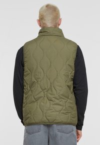 Olivgrüne gesteppte Weste mit hohem Kragen, ärmellosem Design und strukturierter Oberfläche. Tragend über einem schwarzen Langarmshirt.