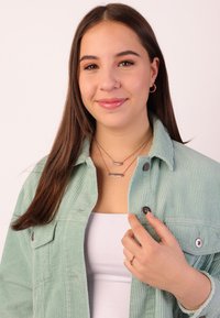 Veste en velours côtelé vert clair avec deux poches poitrine, chaînes en or superposées avec des pendentifs rectangulaires, et un haut blanc en dessous.