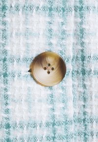 Okrągły, beżowy guzik z czterema otworami, umieszczony na teksturowanej tkaninie w zielono-białą kratę.