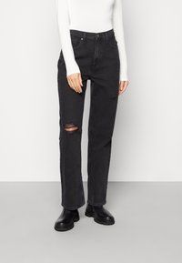 Svarta denimjeans med en avslappnad passform, med slitna detaljer vid knäna. Kombineras med svarta kraftiga stövlar och en vit långärmad topp.