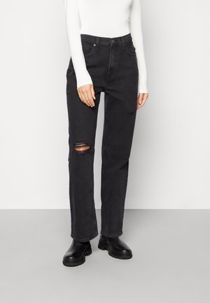 Jeans de mezclilla negra con un ajuste relajado, que presentan un detalle desgastado en la rodilla. Combinados con botas negras gruesas y una camiseta blanca de manga larga.