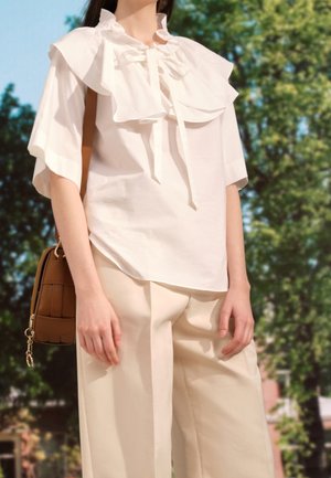 Personne portant un chemisier blanc à volants et un pantalon large crème, portant un sac à bandoulière marron, debout en plein air avec des arbres en arrière-plan.