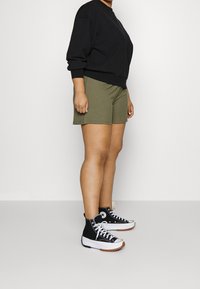 Sweatshirt noir, shorts côtelés vert olive, et baskets montantes noires avec semelles blanches et semelles en caoutchouc naturel. Le modèle se tient de profil.