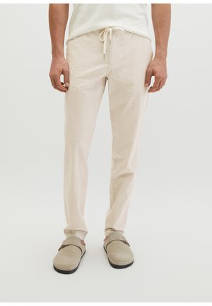 Man draagt beige broek met trekkoord, beige instapschoenen en een wit overhemd, staand tegen een effen lichte achtergrond.