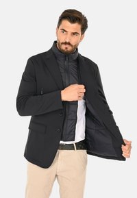 Blazer nero con uno strato interno isolato e con cerniera. Presenta una texture liscia, bottoni e due tasche laterali. Indossato sopra una camicia bianca.