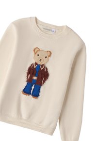 Gehaakte trui in crème met een berenontwerp. Heeft een bruine franjenjack en blauwe spijkerbroek. Geribbelde manchetten en zoom. Zachte textuur.