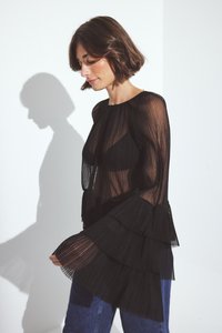 Blusa nera plissettata con maniche lunghe a volant. Tessuto trasparente con un design a strati. Indossata sopra un indumento nero. Silhouette su uno sfondo chiaro.