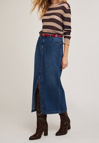 Jupe en denim avec fente devant, présentant un lavage bleu foncé et un design simple. Portée avec un pull en tricot rayé et des bottes à talons hauts en imprimé serpent.