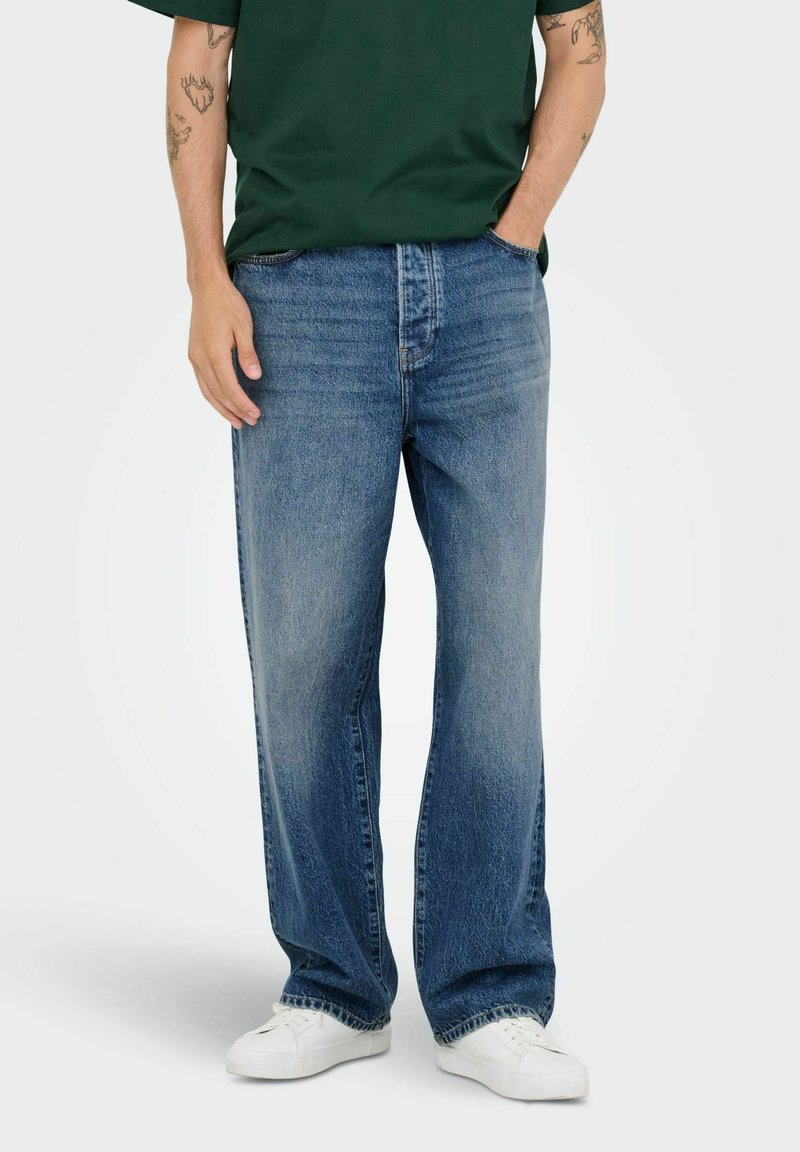 Ljust blå denimjeans med en avslappnad passform, med en klassisk femficksdesign och rak skärning, bärs tillsammans med en grön t-shirt och vita sneakers.