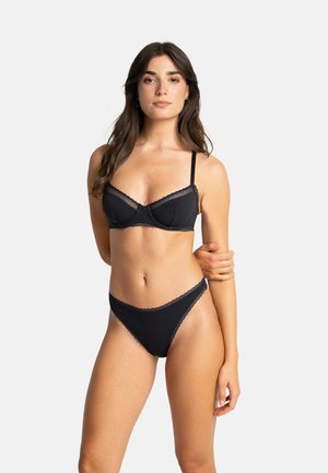 Femme aux longs cheveux foncés portant un soutien-gorge noir bordé de dentelle et une culotte taille haute assortie, debout, un bras croisé sur son corps.