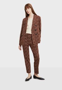 Ruskea kuvioitu puku, jossa on kaksirivinen bleiseri ja slim-fit-housut. Kangas sisältää pyöreitä kuvioita. Yhdistetty vaalean toppauksen ja mustien tasapohjaisten kenkien kanssa.