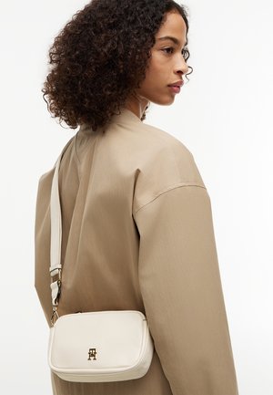 Donna con capelli ricci, giacca beige e una piccola borsa a tracolla bianca con dettagli dorati sulla schiena.