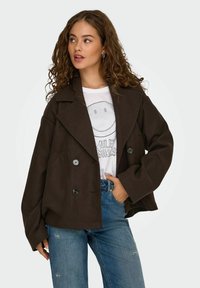 Bruine dubbelzijdige jas met grote revers en zwarte knopen, gecombineerd met een wit graphic t-shirt en lichtblauwe jeans.
