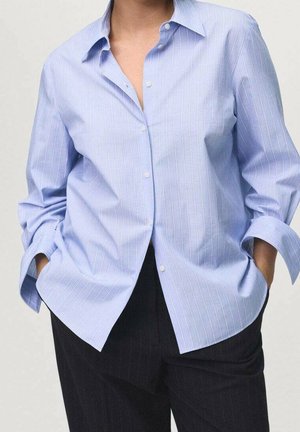 Person iført en lyseblå skjorte med fine striber, knapper og opkørte ærmer samt hænderne i lommerne, kombineret med sorte bukser med fine striber.