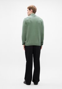 Green sweatshirt with long sleeves, ribbed cuffs, and hem, paired with loose black trousers. The model is viewed from the back against a white background.