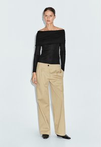 Schwarzes, schulterfreies Langarmtop, kombiniert mit beigen, weiten Hosen mit Falten und sichtbarem Knopfdetail. Schwarze Schuhe.