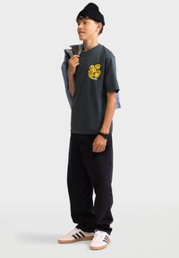 Camiseta de algodón verde oscuro con un gráfico de una cara sonriente amarilla en el pecho, jeans negros y zapatillas blancas con rayas negras.