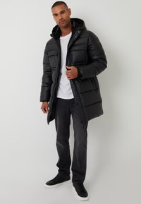 Sort polstret jakke med hætte, der har lynlås- og knappelukning, to frontlommer og en glat, blank overflade. Brugt med grå jeans og sorte sneakers.