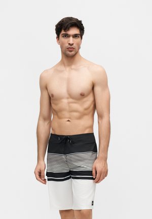 Mężczyzna bez koszulki z krótkimi ciemnymi włosami, ubrany w czarno-białe pasiaste kąpielówki, stojący na tle jednolitego jasnego tła.