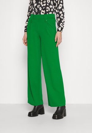 Mujer con pantalones verdes brillantes de talle alto y pierna ancha, botines con plataforma negros y blusa de manga larga con estampado en blanco y negro.