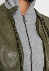 Olivgrön läderjacka med tryckknappar i kragen, ozipad över en grå hoodie med framzip och texturerat tyg.