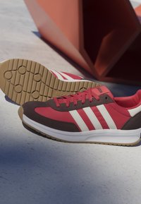 Rote und braune Adidas-Sneaker mit weißen Streifen und Gummisohlen, platziert auf einer hellen Oberfläche mit einer roten geometrischen Form im Hintergrund.