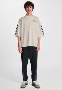 Beige oversize-t-shirt med svart logotyp och svarta diagonala ränder på ärmarna; matchad med svarta byxor och beige-grå sneakers.