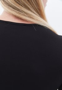 Schwarzes Oberteil mit rundem Ausschnitt, glatter Textur und ohne sichtbare Muster oder Verzierungen. Haare verdecken teilweise den hinteren Ausschnitt.