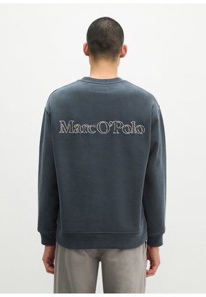 Persoon gezien van achteren die een donkerblauwe trui draagt met de tekst "Marc O'Polo" geborduurd over de bovenrug.