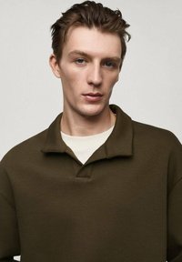 Polo shirt oversize di colore verde oliva, realizzato in tessuto morbido, con colletto e patta con bottoni a metà. Indossato sopra una camicia chiara.