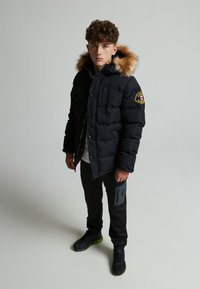 Black puffer jacket with a detachable fur-lined hood, prominent chest pocket, and patch accents, paired with black pants featuring a zip pocket.