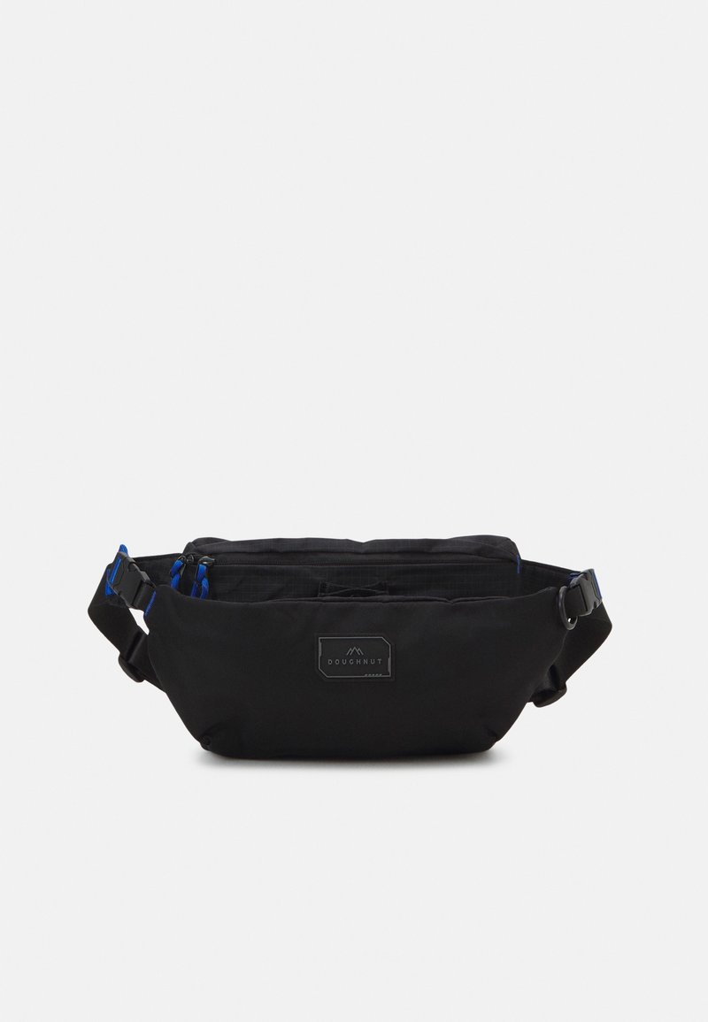 Sac banane noir avec sangle ajustable, deux compartiments zippés avec tirettes bleues, et un patch rectangulaire marqué « Doughnut » à l'avant.