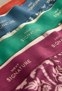 Quatre ceintures élastiques colorées en orange, bleu, vert et violet, chacune avec le texte "NEXT SIGNATURE" en lettres dorées.