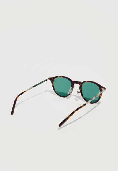 Στρογγυλά γυαλιά ηλίου με σκελετούς από φίδι, φακούς με πράσινη απόχρωση και ασημένιους μεταλλικούς βραχίονες πάνω σε λευκό φόντο.