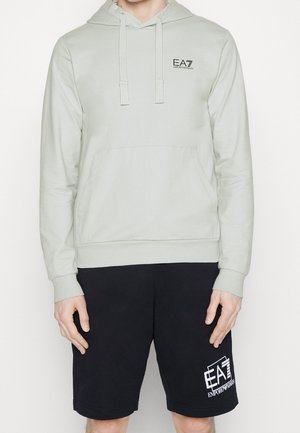 Persona che indossa una felpa con cappuccio grigio chiaro EA7 Emporio Armani e pantaloncini neri EA7 Emporio Armani, in piedi davanti a uno sfondo bianco neutro.