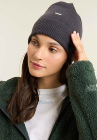 Beanie grigio a maglia con una texture a coste e un accento argentato. Indossato con una giacca verde in pelliccia e una t-shirt bianca sotto.
