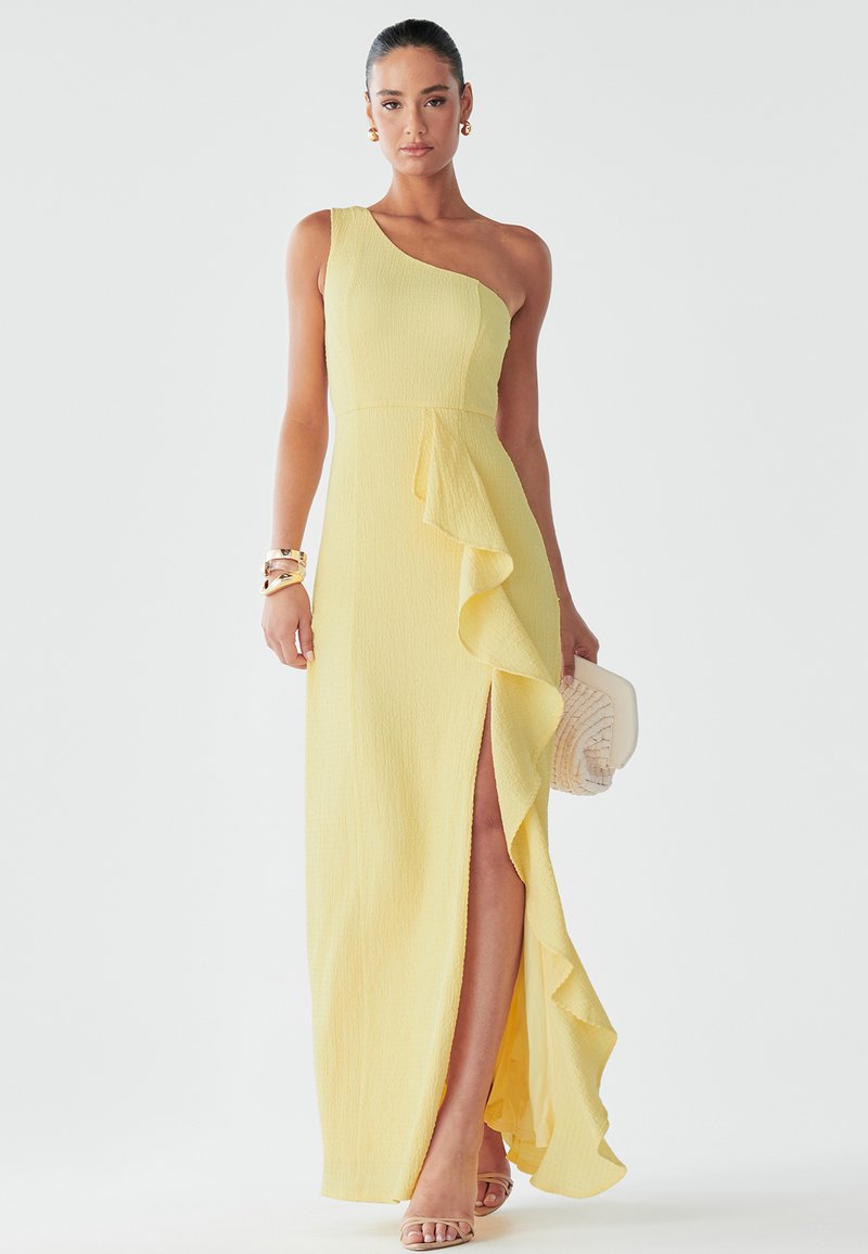 Asymmetrical yellow dress with textured fabric, ruffle detail on the side, and a thigh-high slit. Worn with a clutch and sandals.