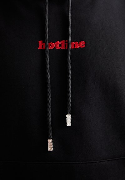 Črna kapuca z rdečim vezenjem "hotline". Vključuje vrvi z kovinskimi konicami in mehko, teksturirano tkanino.