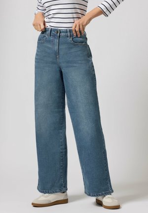 Osoba ubrana w jasnoniebieskie szerokie dżinsy, zapinająca pasek, w zestawieniu z białymi tenisówkami i białym topem z czarnymi poziomymi paskami.