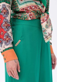 Groene broek gemaakt van gladde stof, met een elastische tailleband. Een blouse met een meerkleurig patroon heeft oranje manchetten, en een logolabel is zichtbaar.