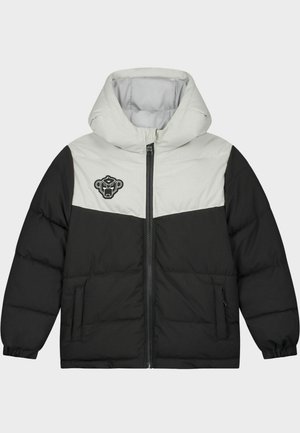 Enfant portant une doudoune à capuche noire et beige, un pantalon de jogging noir et des baskets blanches, debout devant un fond blanc.