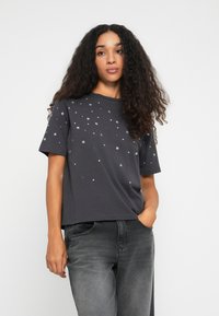 Ung kvinde med langt krøllet hår iført en mørkegrå t-shirt med hvide stjernesyn og slidte sorte jeans, står med den ene hånd i lommen.