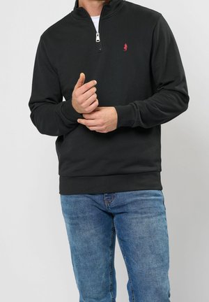 Mężczyzna w czarnej bluzie z suwakiem do połowy i małym czerwonym logo na piersi oraz niebieskich dżinsach, poprawia lewe nadgarstek prawą ręką.