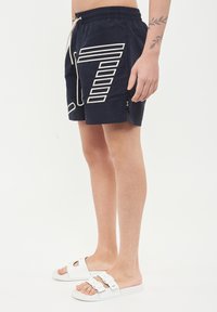 Mjärta simbyxor i marinblått med elastisk midja och dragsko, med vitt geometriskt mönster på sidan och kontrasterande dragkedjeficka.