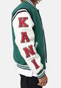 Giacca varsity verde con maniche bianche. Presenta la scritta ricamata rossa "KANI." Include colletto e polsini a righe a costine.