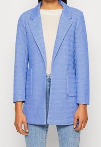 Blazer azul de espiga con solapas de muesca, mangas largas y dos bolsillos delanteros, confeccionado en una tela texturizada. Llevado sobre una blusa clara y jeans.