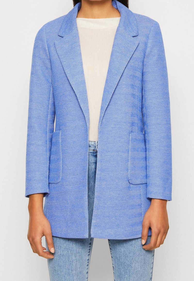 Blazer azul de espiga con solapas de muesca, mangas largas y dos bolsillos delanteros, confeccionado en una tela texturizada. Llevado sobre una blusa clara y jeans.